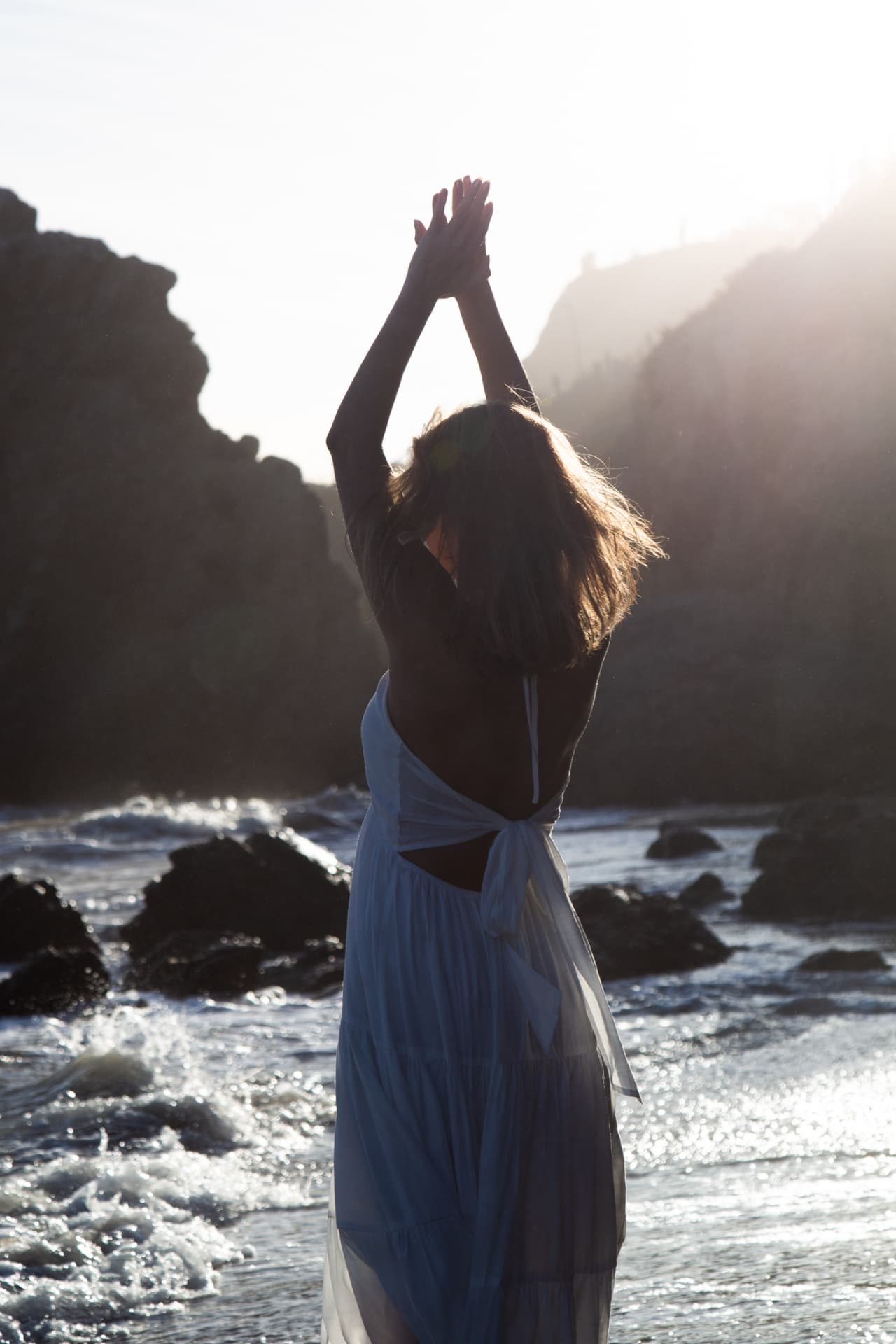 A woman standing in the ocean at sunset, arms raised toward the light